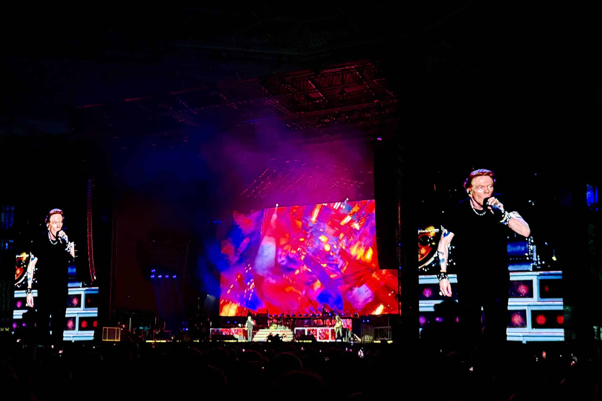 Cantor vestido de preto canta no centro do palco durante show noturno. Telões grandes exibem imagens coloridas e ampliam a figura do artista. Público está em frente ao palco, visível em silhueta.
