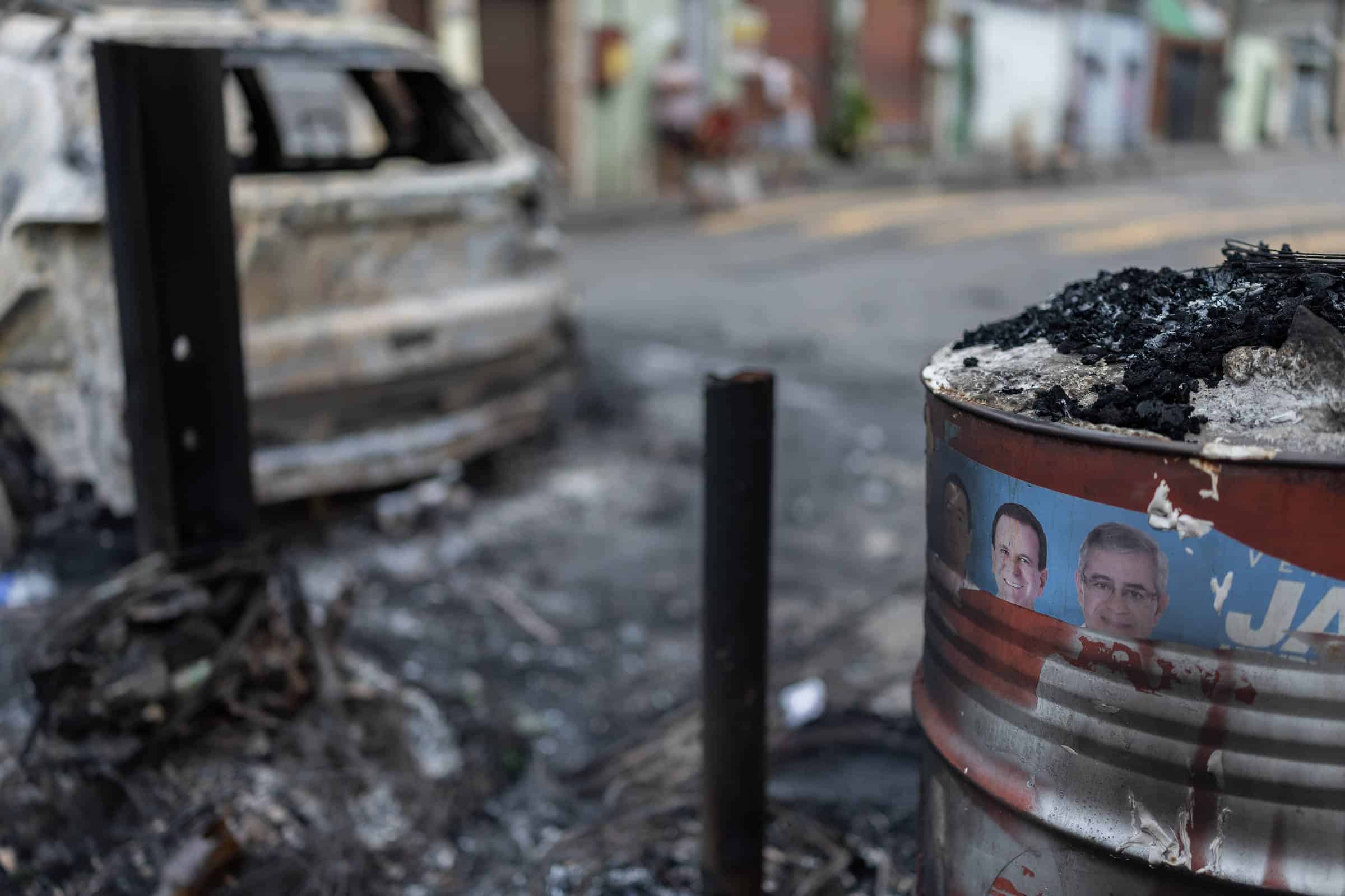 Carro completamente queimado estacionado em rua com calçada e casas ao fundo. Em primeiro plano, barril metálico com cinzas e carvão, exibindo adesivo com rostos de três homens. Ambiente mostra sinais de incêndio recente.