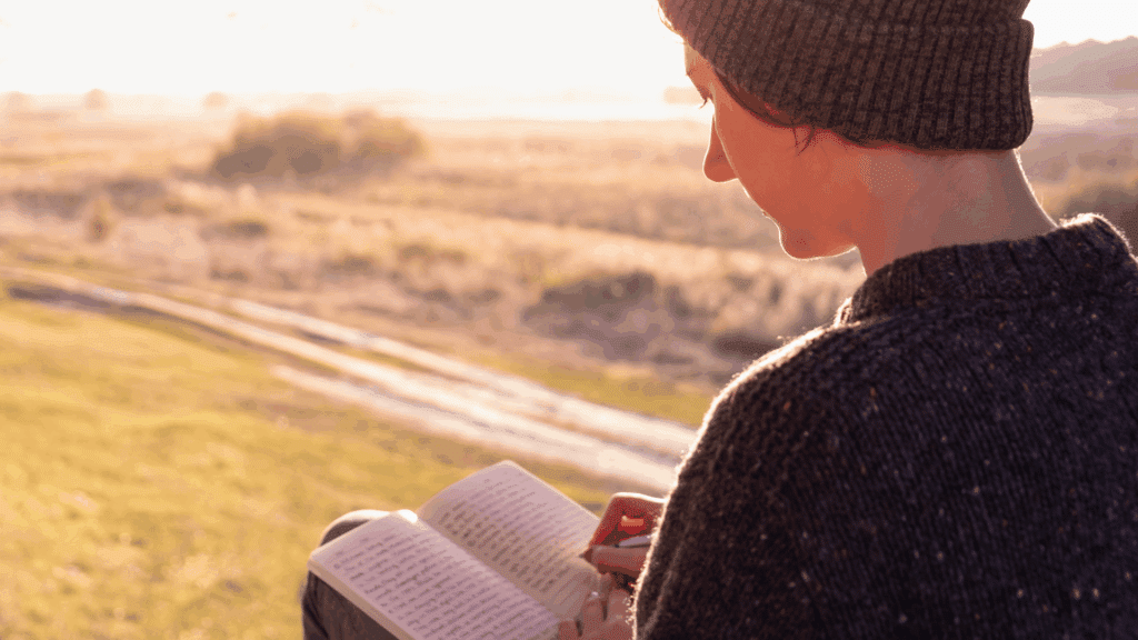 Uma mulher está sentada ao ar livre e em um ambiente de natureza. Ela está escrevendo em um diário ou caderno. Ao fundo, há o pôr do sol.