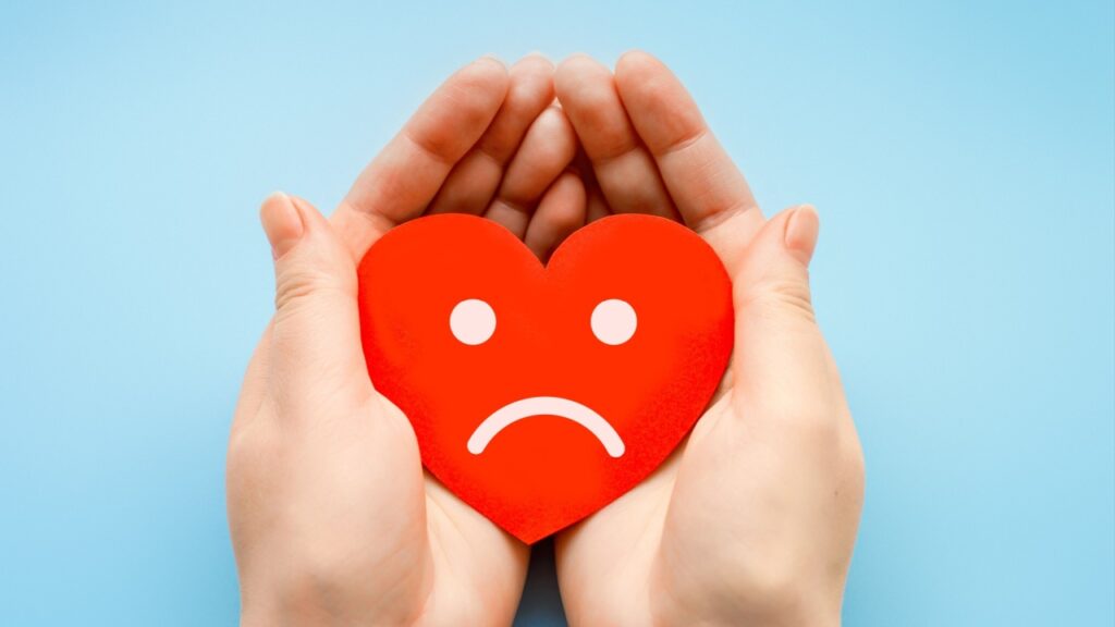 coração com uma carinha triste na cor branca na palma de um par de mãos em fundo azul