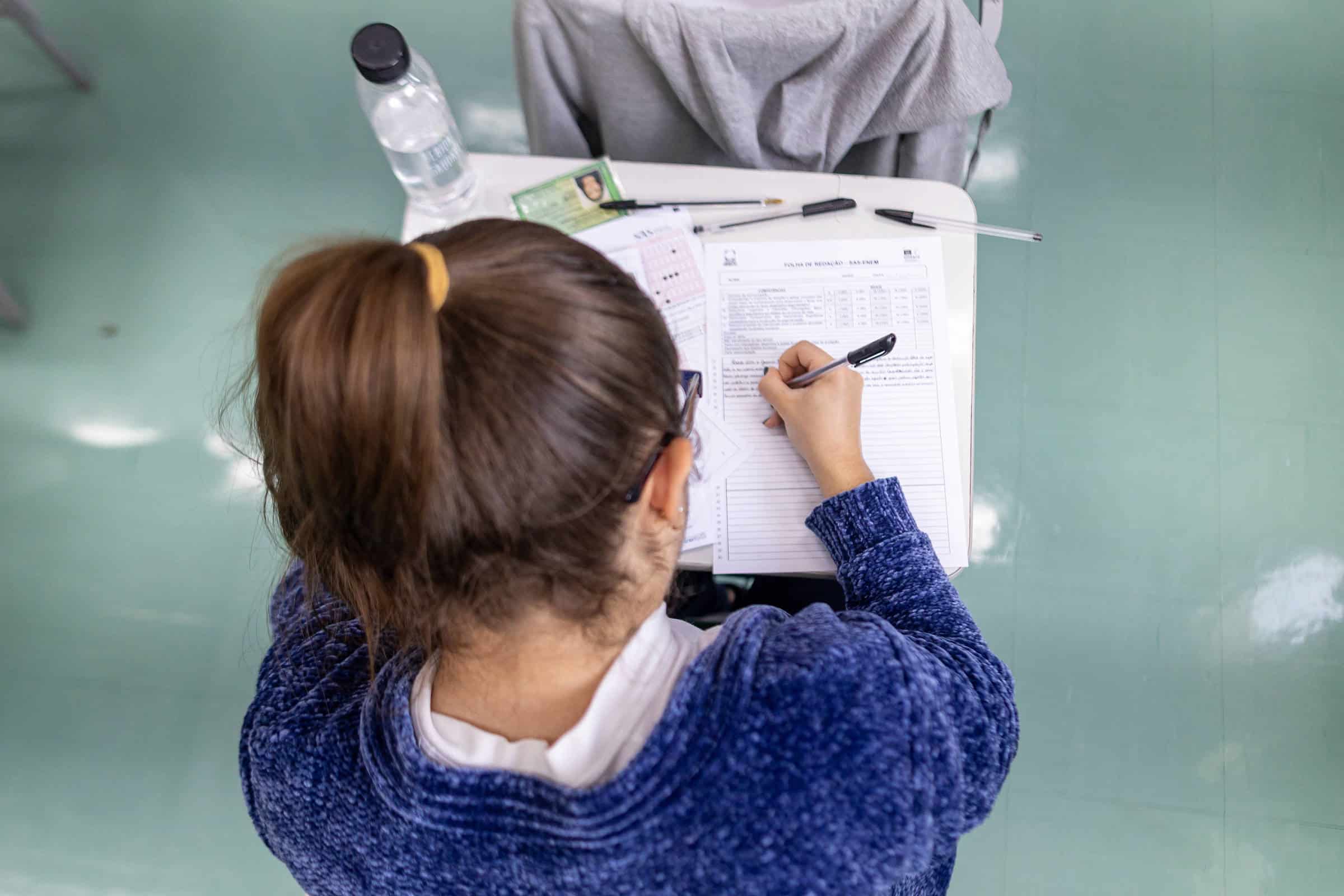 A imagem mostra a parte de trás de uma estudante com cabelo preso em um coque, usando um suéter azul. Ela está escrevendo em uma folha de papel sobre uma mesa, que também contém uma garrafa de água, canetas e outros papéis. O ambiente parece ser uma sala de aula, com piso verde.