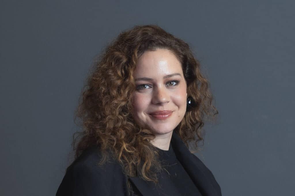 Mulher de cabelos cacheados e castanhos posa de perfil com leve sorriso, vestindo blazer preto sobre blusa preta, com fundo cinza neutro.