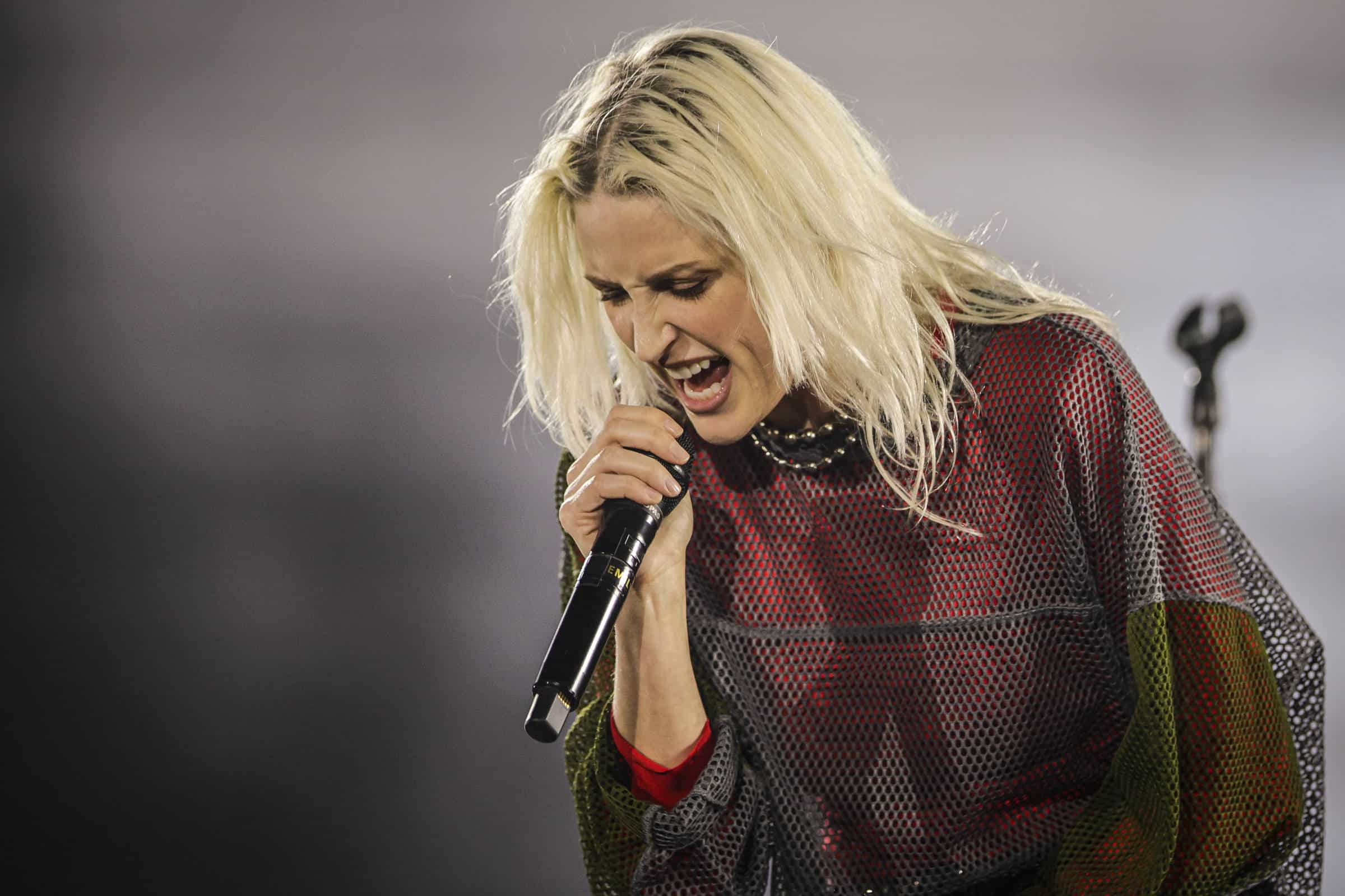 Mulher loira com cabelo solto segura microfone próximo à boca enquanto canta intensamente. Ela veste blusa de malha preta com detalhes vermelhos e verdes, em fundo desfocado.