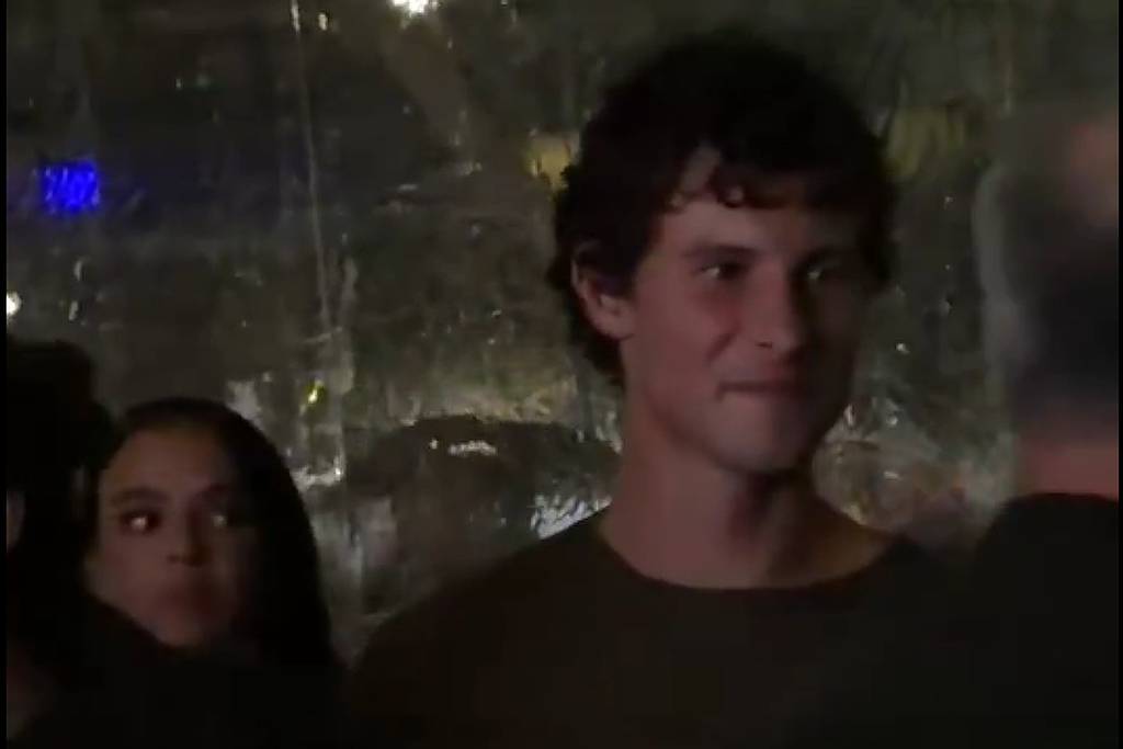 Homem jovem de cabelos curtos e escuros veste camiseta escura, está em ambiente com iluminação baixa e fundo desfocado. Ao lado esquerdo, mulher com cabelo longo e escuro observa algo fora do quadro.