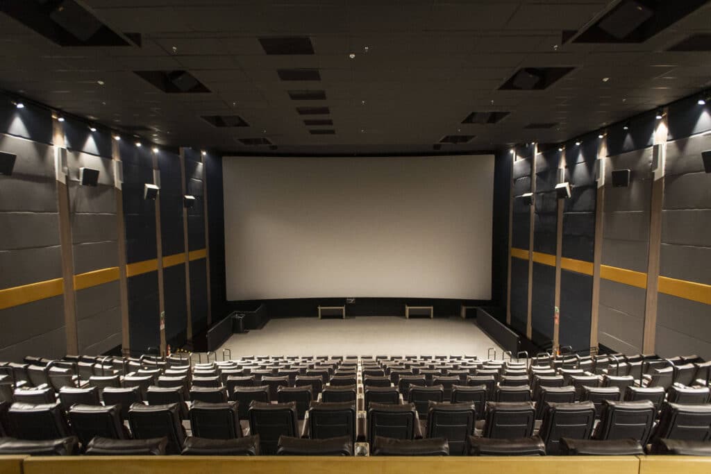 Sala de cinema com fileiras de poltronas pretas vazias voltadas para uma grande tela branca. As paredes laterais são cinza com faixas amarelas e iluminação embutida no teto e nas paredes.