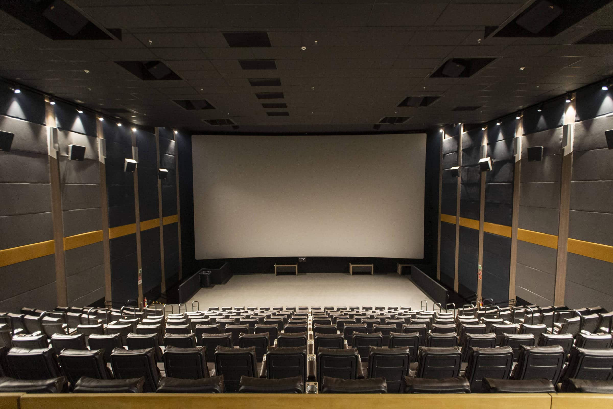 Sala de cinema com fileiras de poltronas pretas vazias voltadas para uma grande tela branca. As paredes laterais são cinza com faixas amarelas e iluminação embutida no teto e nas paredes.