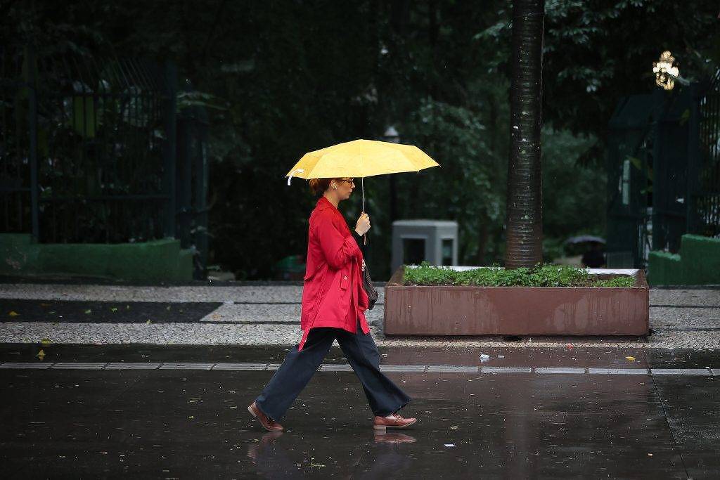 Pessoa vestindo casaco vermelho caminha na calçada molhada segurando um guarda-chuva amarelo aberto. Ambiente urbano com árvores e bancos ao fundo em dia escuro e chuvoso.