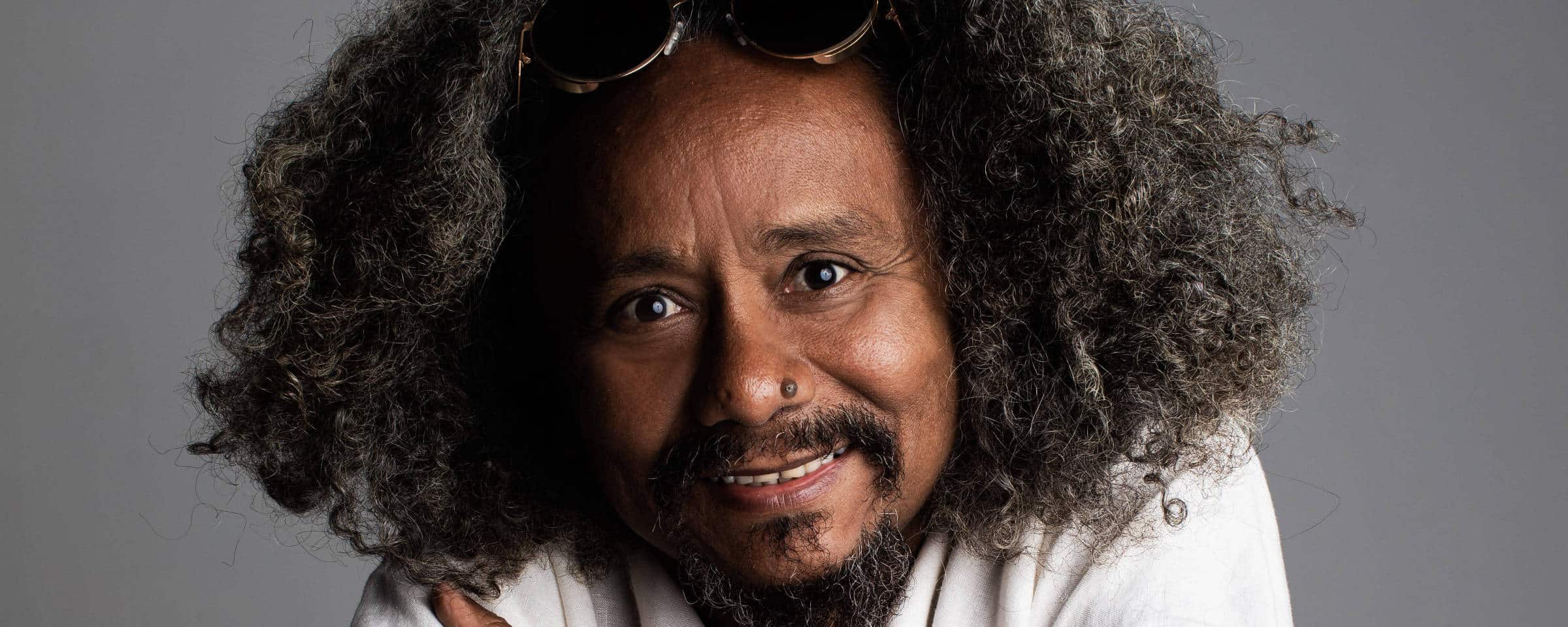 Homem de meia-idade com cabelo afro volumoso e barba, usando camisa branca, sentado de costas em cadeira vermelha, apoiando os braços no encosto.