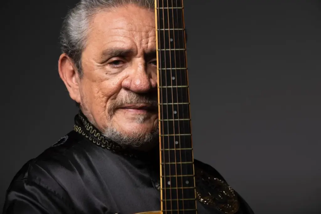 A imagem mostra um homem mais velho, com cabelos grisalhos e barba rala, vestido com uma camisa preta de tecido brilhante com detalhes dourados no colarinho. Ele está segurando um violão na posição vertical, de forma que o braço do instrumento divide seu rosto ao meio. O fundo é neutro e escuro, o que ajuda a destacar o sujeito da foto. O olhar do homem é sereno e transmite uma expressão de sabedoria e tranquilidade. A composição da imagem, com o violão centralizado entre o rosto dele, sugere uma forte ligação com a música, talvez indicando que ele é músico, cantor ou compositor. A iluminação é suave e bem direcionada, realçando as texturas da pele e do instrumento. É um retrato artístico e intimista.