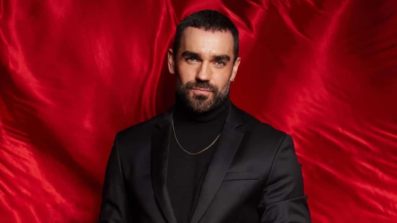 Marcus Ornellas de roupa social preta, olhando sério para a câmera, posando em fundo vermelho de pano