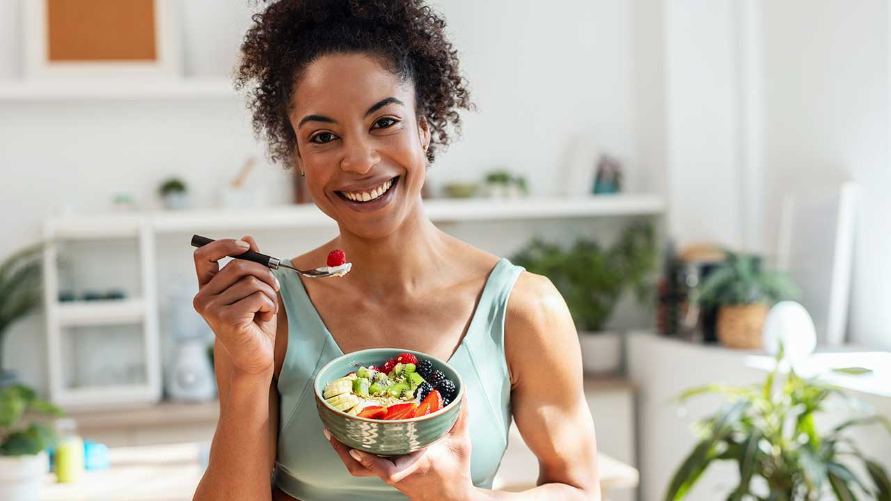 mulher negra segurando um garfo e segurando uma cumbuca com frutas em uma cozinha