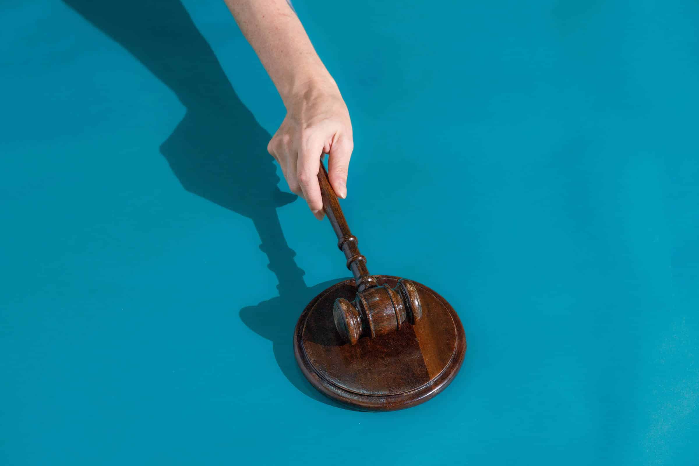 Mão segura martelo de juiz de madeira sobre base redonda de madeira, fundo azul.