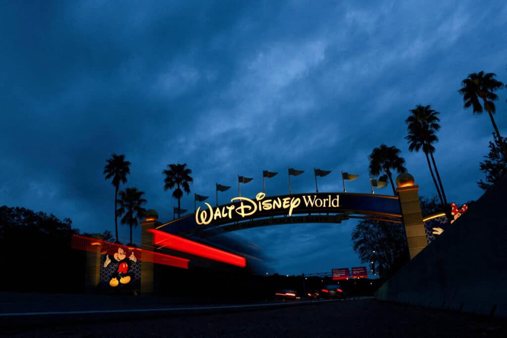A imagem mostra o arco de entrada do Walt Disney World, iluminado em meio a um céu nublado e escuro. O arco é decorado com o nome