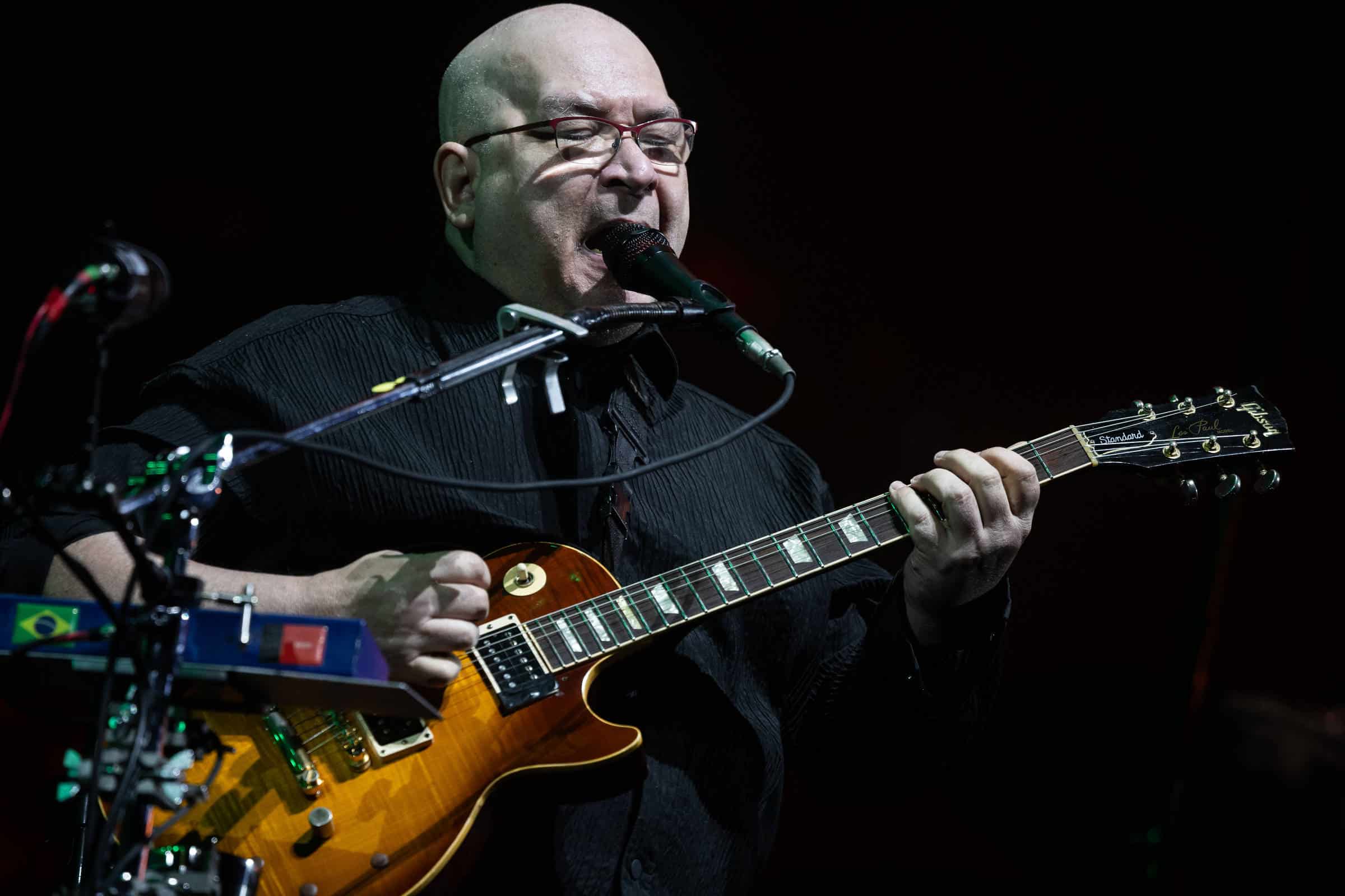 Um homem calvo, usando óculos e uma camisa preta, está tocando guitarra em um palco. Ele canta em um microfone e tem um equipamento musical ao seu redor. A guitarra é de cor dourada e ele parece concentrado na performance.