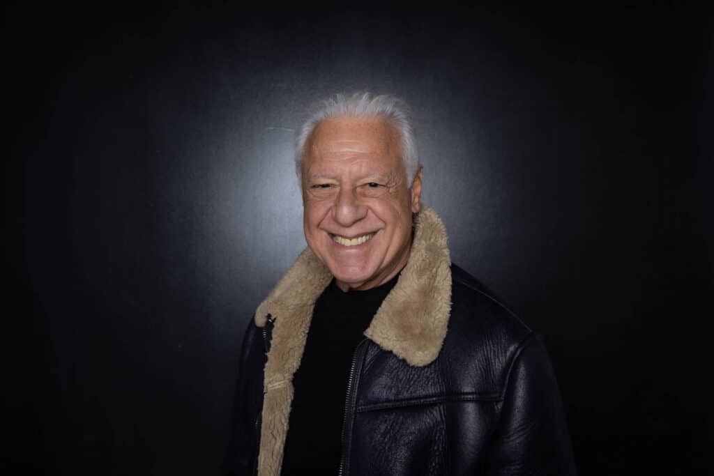 Um homem sorridente está posando para a foto. Ele tem cabelo grisalho e usa uma jaqueta de couro escura com um colarinho de pelagem clara. O fundo da imagem é preto, destacando o rosto do homem.