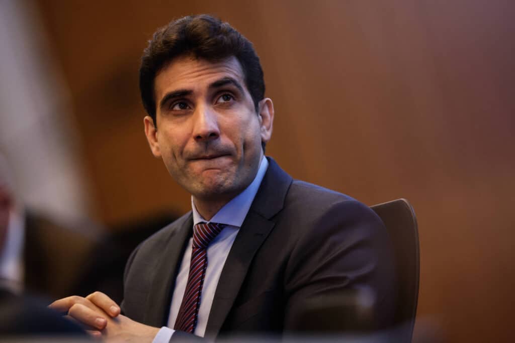 Um homem com cabelo escuro e liso, usando um terno escuro e uma gravata listrada, está sentado em uma mesa. Ele parece pensativo, com a boca levemente entreaberta e as mãos unidas na frente. O fundo é desfocado, sugerindo um ambiente de reunião ou conferência.