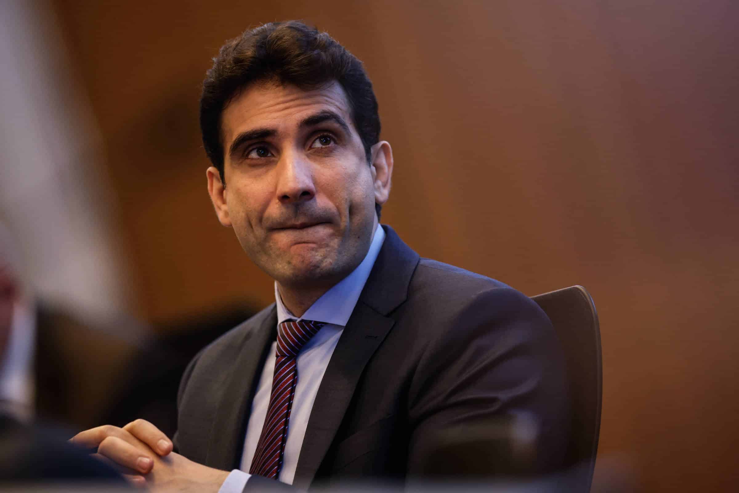 Um homem com cabelo escuro e liso, usando um terno escuro e uma gravata listrada, está sentado em uma mesa. Ele parece pensativo, com a boca levemente entreaberta e as mãos unidas na frente. O fundo é desfocado, sugerindo um ambiente de reunião ou conferência.