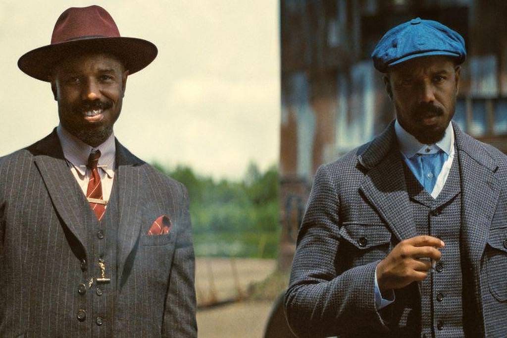 Dois homens posam lado a lado. O da esquerda usa terno risca de giz cinza escuro, gravata vermelha com padrão, colete, lenço no bolso e chapéu vinho. O da direita veste terno cinza com colete, camisa azul clara, gravata azul escura e boina azul. Fundo desfocado sugere área externa.