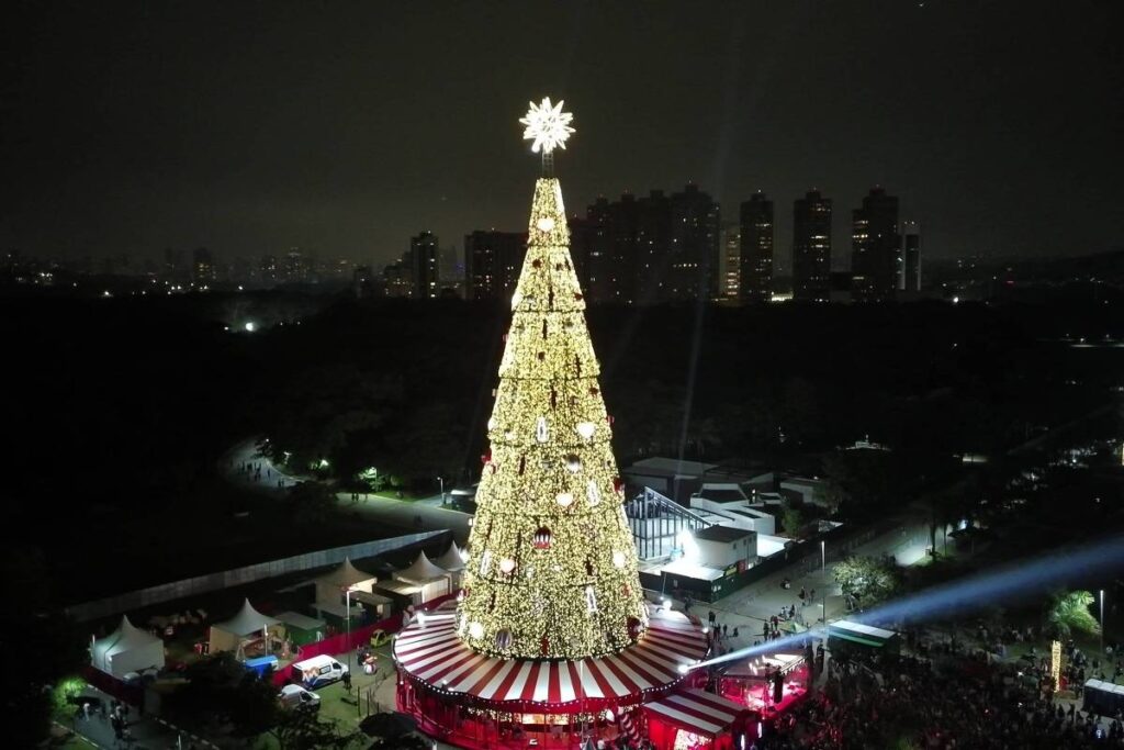 Árvore de Natal gigante decorada com luzes douradas e estrela no topo, situada em praça com tenda vermelha e branca na base. Multidão observa e circula ao redor durante a noite, com prédios ao fundo e iluminação pontual no ambiente.