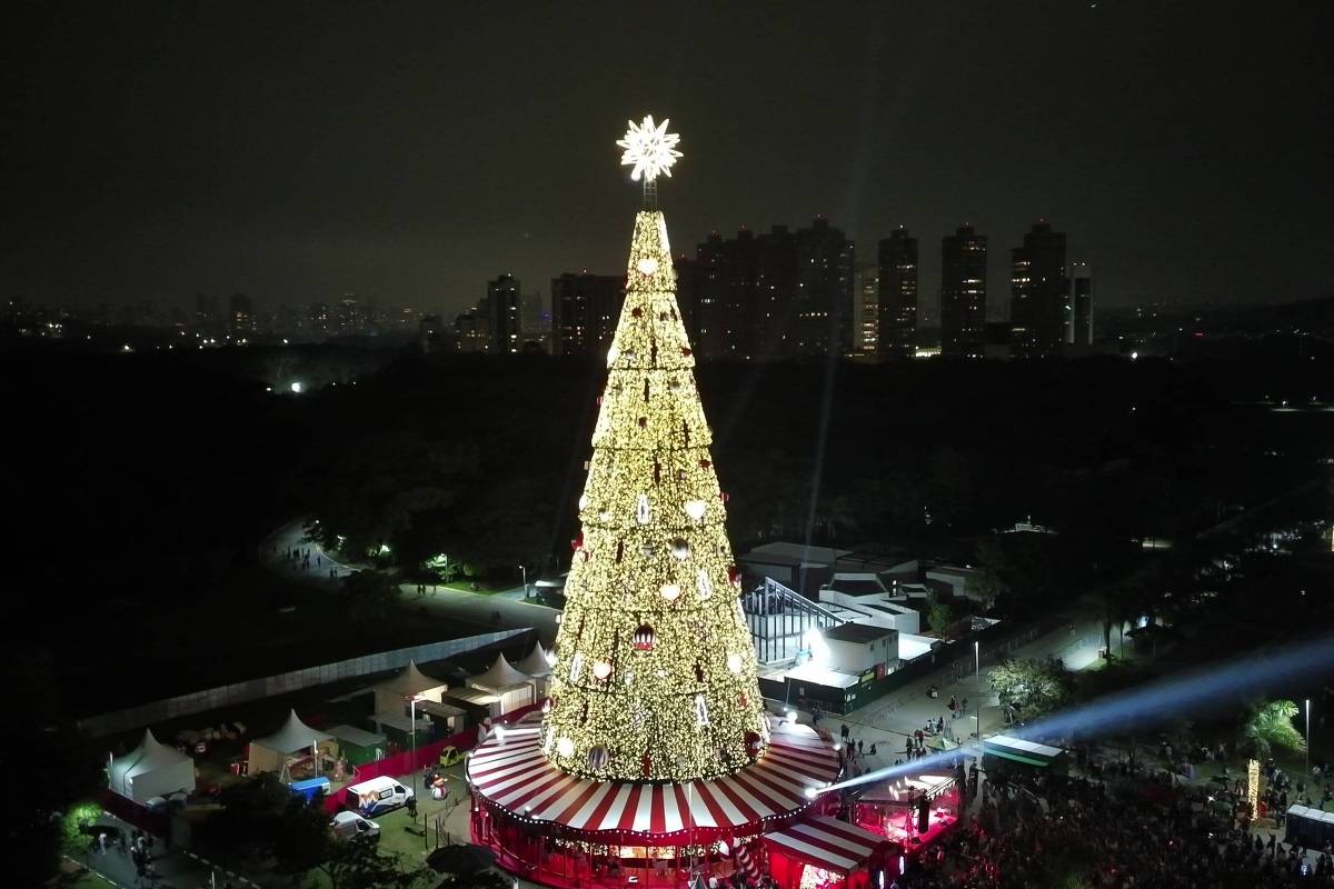 Árvore de Natal gigante decorada com luzes douradas e estrela no topo, situada em praça com tenda vermelha e branca na base. Multidão observa e circula ao redor durante a noite, com prédios ao fundo e iluminação pontual no ambiente.