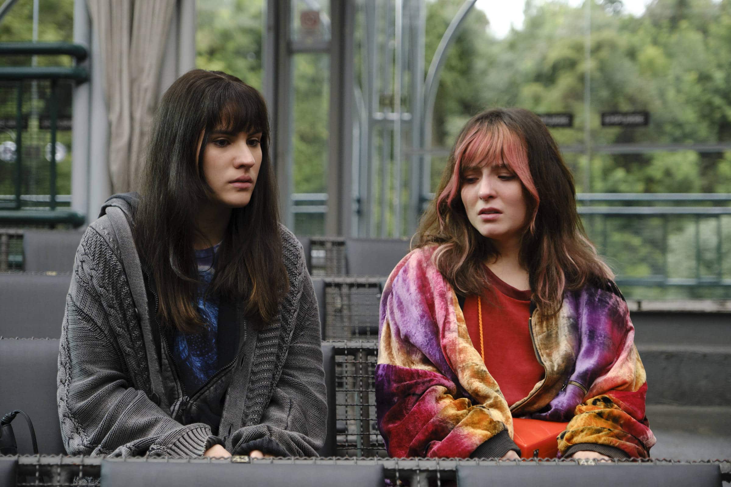 Duas mulheres sentadas lado a lado em cadeiras de vime em um ambiente externo coberto. A mulher à esquerda veste suéter cinza e camiseta azul, com cabelo castanho escuro e liso. A mulher à direita usa casaco colorido em tons de roxo, laranja e amarelo sobre blusa vermelha, com cabelo castanho claro e ondulado. Ao fundo, vegetação e estrutura metálica visível.