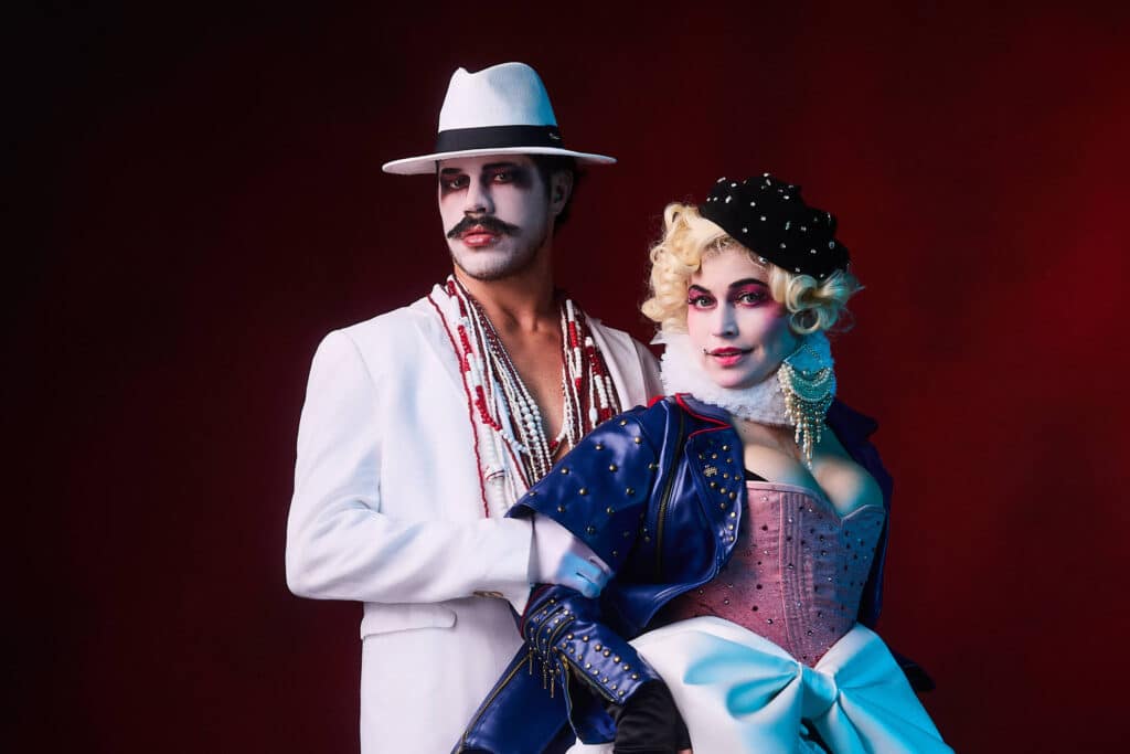 Homem e mulher posam juntos contra fundo vermelho. Ele veste terno branco, chapéu e maquiagem facial, ela usa vestido com corpete rosa, jaqueta azul, chapéu preto com detalhes e maquiagem teatral, levantando uma perna com meia verde e sapato preto.