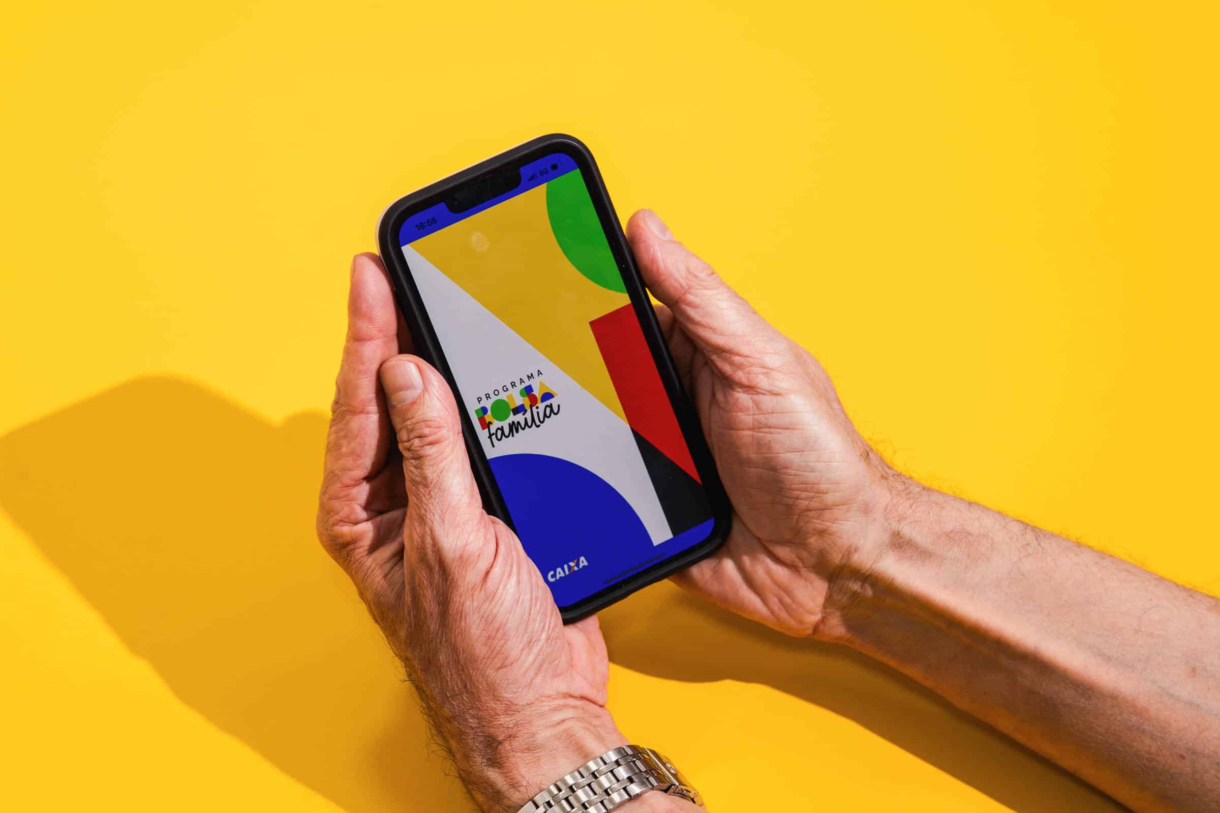 Mãos seguram um celular com tela exibindo formas geométricas coloridas em fundo amarelo. A sombra das mãos e do aparelho aparece à esquerda.