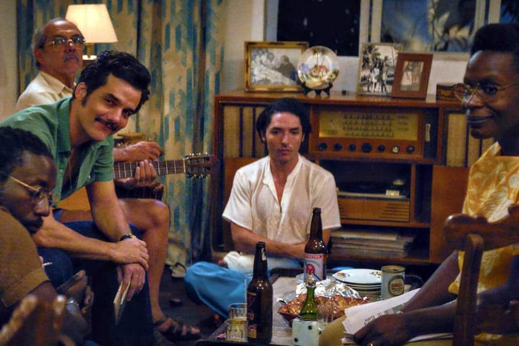 A imagem mostra um grupo de seis pessoas sentadas em um ambiente interno, possivelmente uma sala de estar. Há uma mesa baixa no centro com algumas bebidas e petiscos. As pessoas estão em diferentes posturas, algumas olhando para a câmera e outras em conversação. O ambiente é iluminado de forma suave, com uma decoração simples e um aparelho de som visível ao fundo.