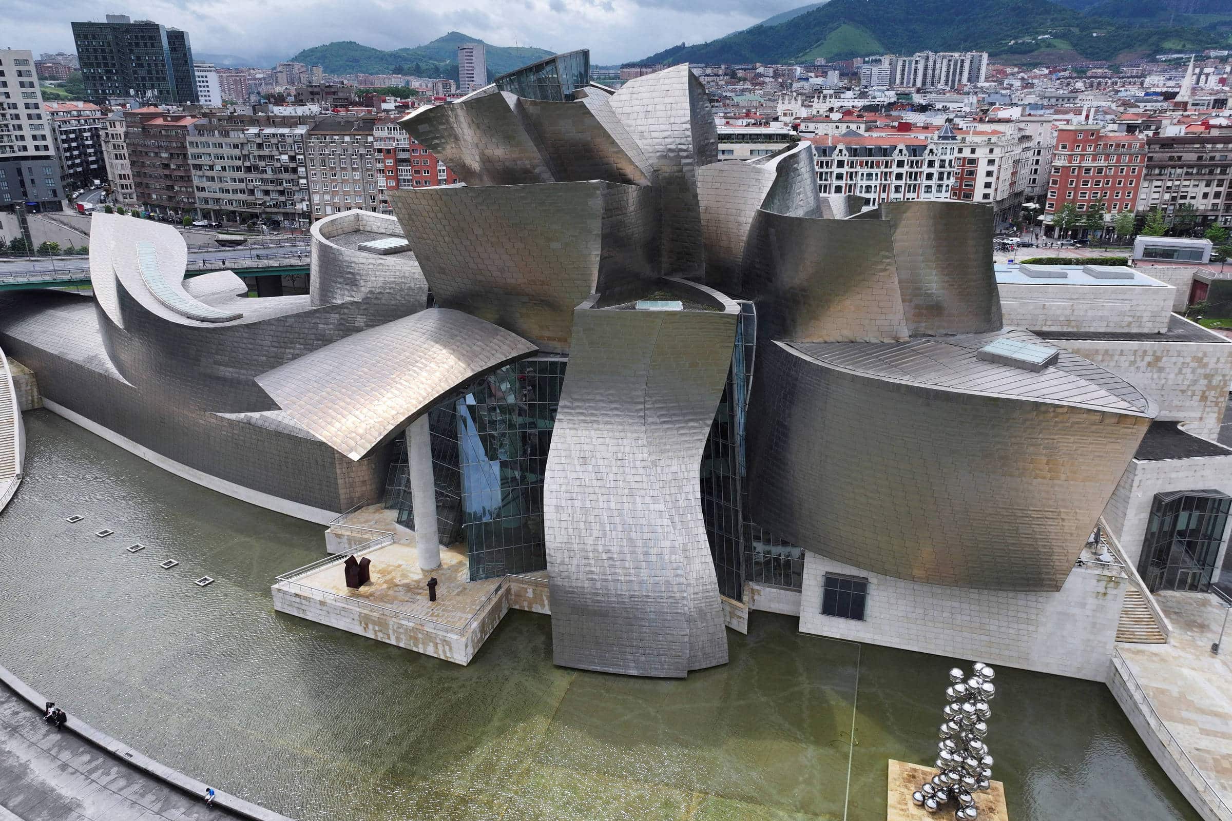 Vista aérea do Museu Guggenheim Bilbao com suas formas curvas e superfícies metálicas refletindo luz. O edifício está cercado por um espelho d