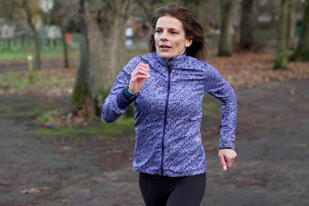 Mulher vestindo jaqueta azul com estampa e calça preta corre em caminho de terra em parque com árvores e folhas secas ao redor.