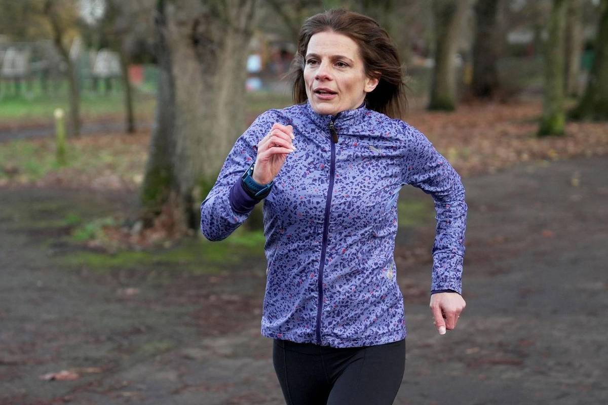 Mulher vestindo jaqueta azul com estampa e calça preta corre em caminho de terra em parque com árvores e folhas secas ao redor.