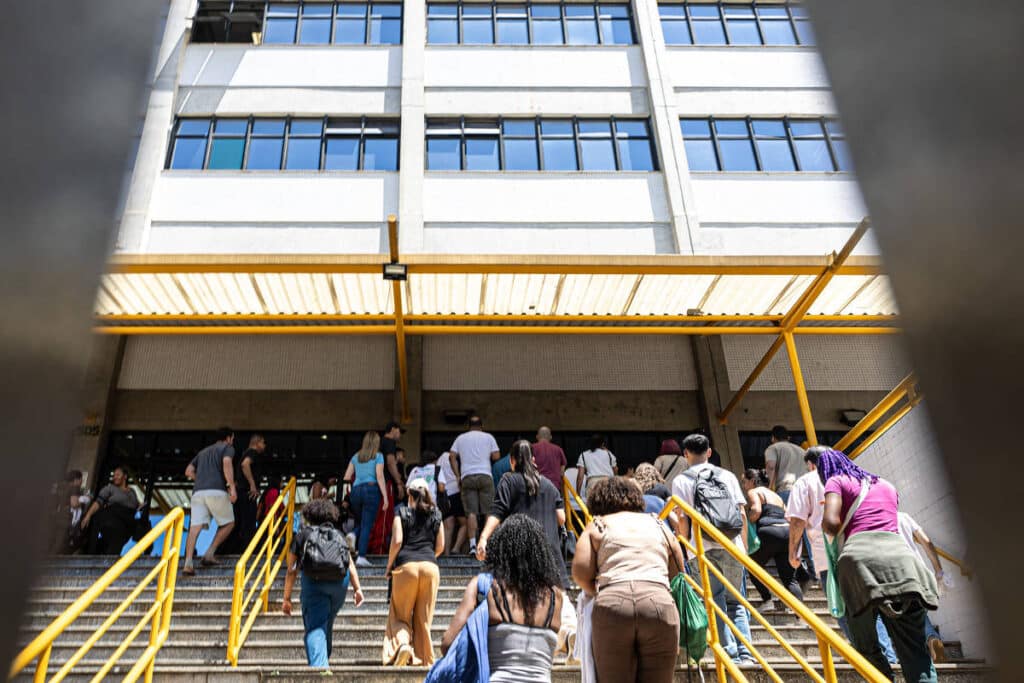 Grupo de pessoas de costas sobe escada com corrimão amarelo em direção à entrada de prédio branco de vários andares com janelas azuis. Estrutura metálica amarela cobre parte da escada.