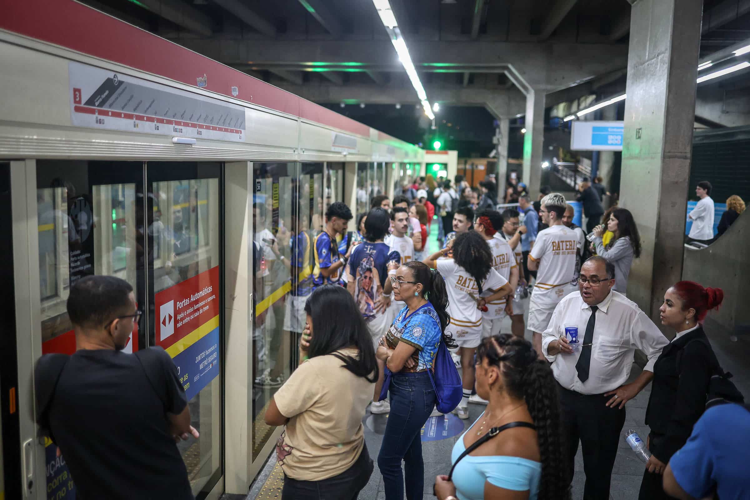 Diversas pessoas aguardam na plataforma para embarcar em trem do metrô. O vagão está parado com portas fechadas e há sinalização visível na lateral. A estação é coberta, com iluminação artificial e colunas de concreto.