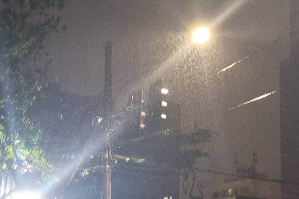 Fumaça densa sai de um prédio residencial à noite, iluminado por luz forte na parte superior. Bombeiros estão no local com caminhão e mangueiras, combatendo o incêndio.