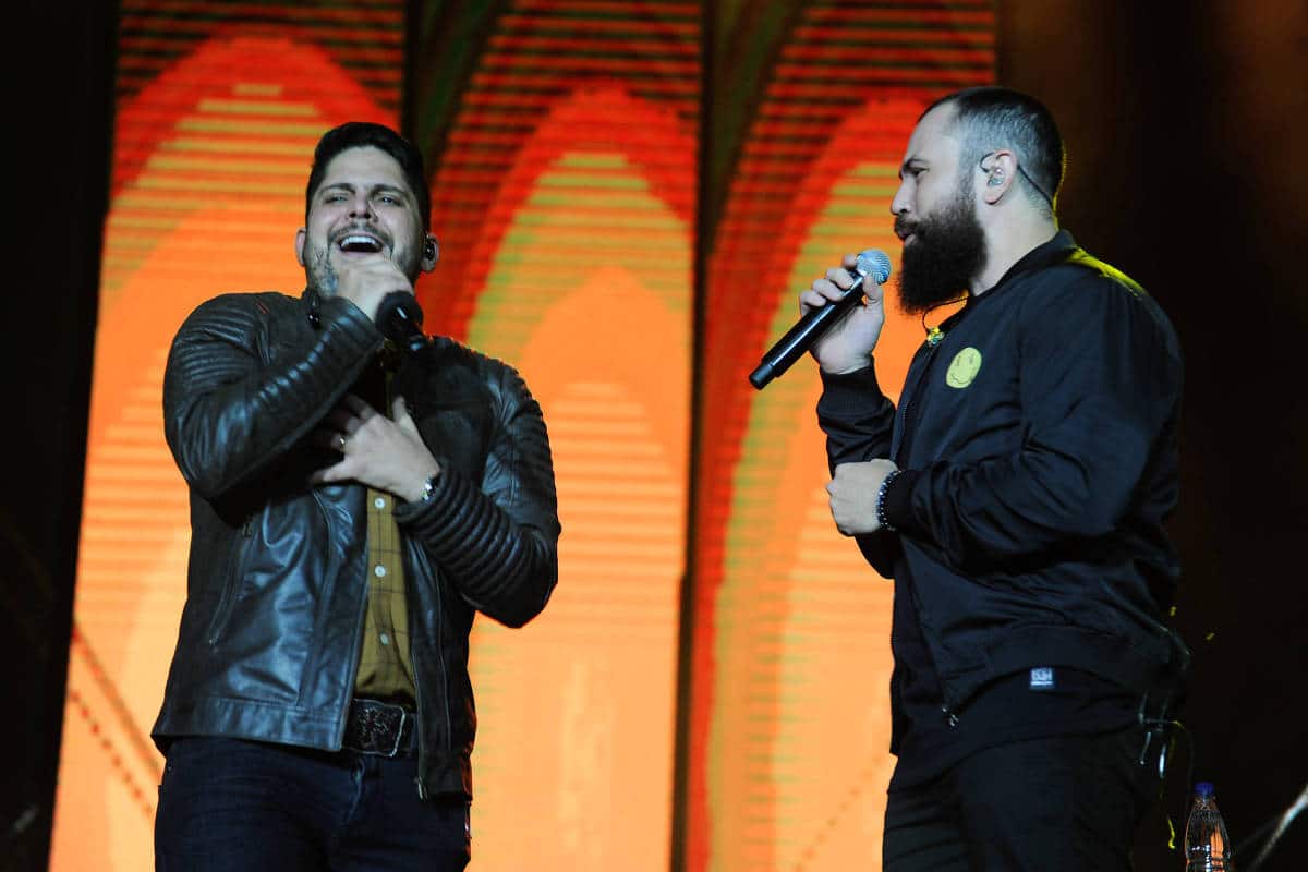 Dois homens cantam no palco, um com jaqueta de couro preta e boné, o outro com jaqueta preta e microfone na mão. Fundo iluminado em tons de laranja e vermelho.