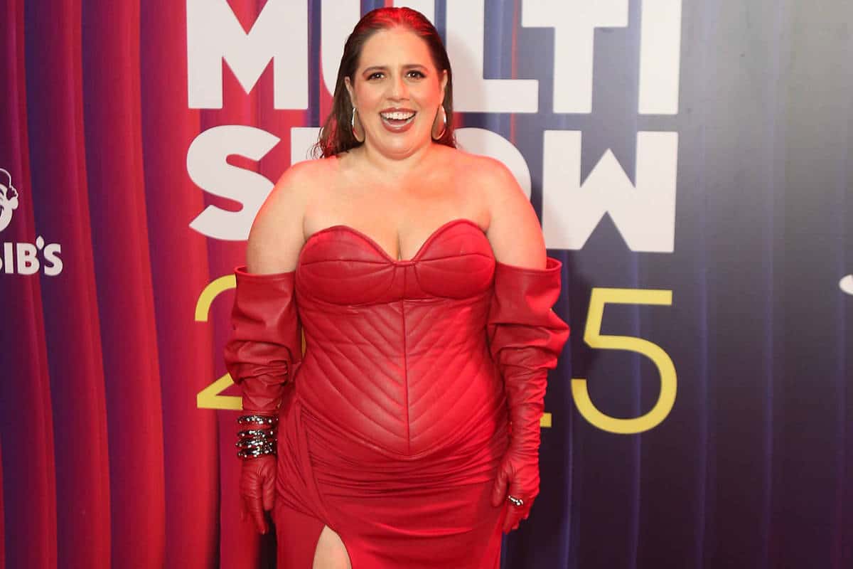Mulher posa no tapete vermelho vestindo vestido longo vermelho com decote coração, mangas bufantes e fenda na perna esquerda. Fundo exibe logo do Prêmio Multishow 2023 em letras amarelas e brancas sobre painel azul e vermelho.