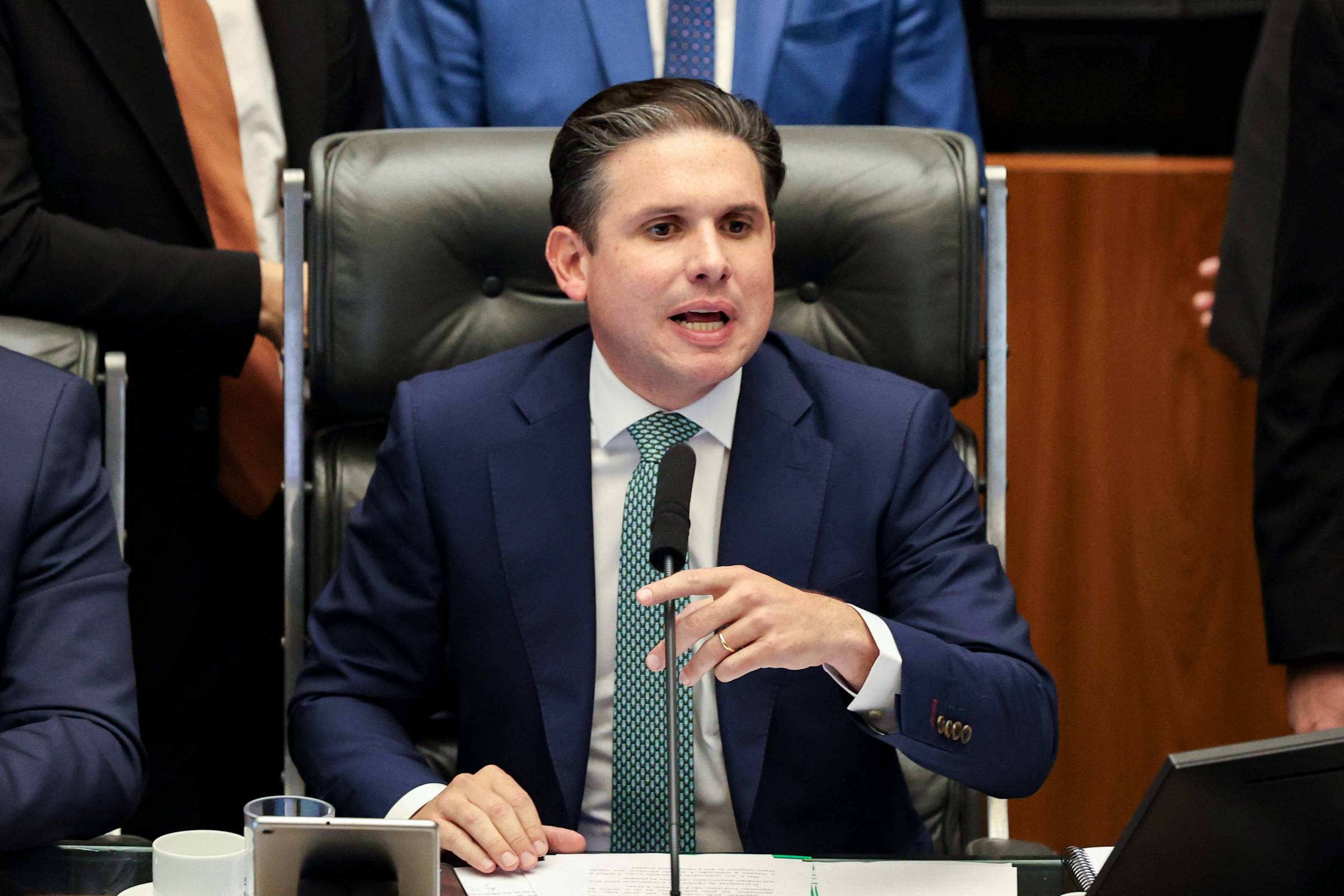 Homem de terno azul escuro e gravata verde senta em cadeira de couro preta, falando ao microfone em ambiente formal, com outras pessoas ao fundo.