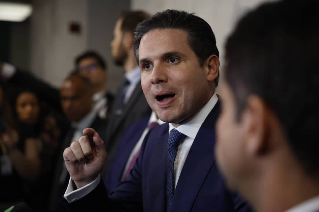 Homem de terno azul e gravata azul fala gesticulando com a mão direita em ambiente interno com outras pessoas ao fundo.