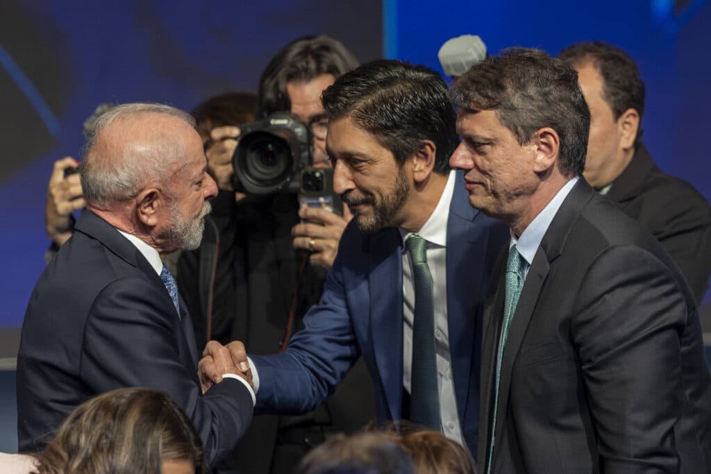 Dois homens de terno apertam as mãos em cumprimento durante evento formal. Um deles tem cabelo grisalho e barba branca, o outro tem cabelo escuro e barba curta. Ao fundo, fotógrafos registram o momento, e há outras pessoas em trajes formais.