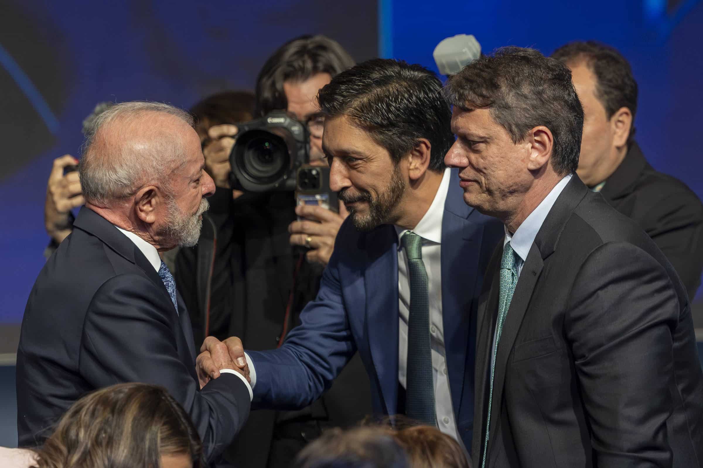 Dois homens de terno apertam as mãos em cumprimento durante evento formal. Um deles tem cabelo grisalho e barba branca, o outro tem cabelo escuro e barba curta. Ao fundo, fotógrafos registram o momento, e há outras pessoas em trajes formais.