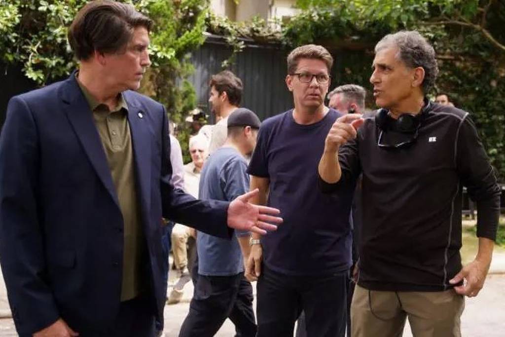Três homens estão em conversa em área externa com árvores e arbustos ao fundo. Um homem à esquerda veste blazer azul e camisa verde, gesticulando com a mão direita. O homem ao centro usa camiseta azul escura e óculos, observando a interação. O homem à direita, com camiseta preta e óculos pendurados no pescoço, gesticula com a mão direita enquanto fala.