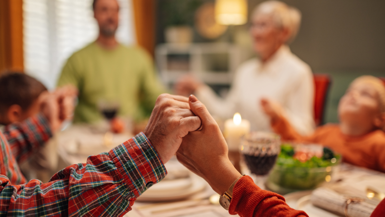 Duas mãos dadas numa mesa de refeição, ao fundo, uma família reunida, com adultos e crianças em clima acolhedor.