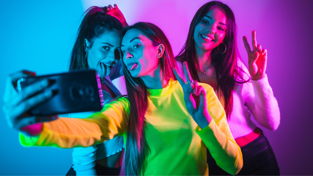Três mulheres sorrindo juntas para uma selfie, fazendo gestos de paz. A mulher da frente segura o celular, enquanto as outras duas mostram descontração. A cena é iluminada com cores azul, rosa e roxo, transmitindo um clima jovem e festivo.
