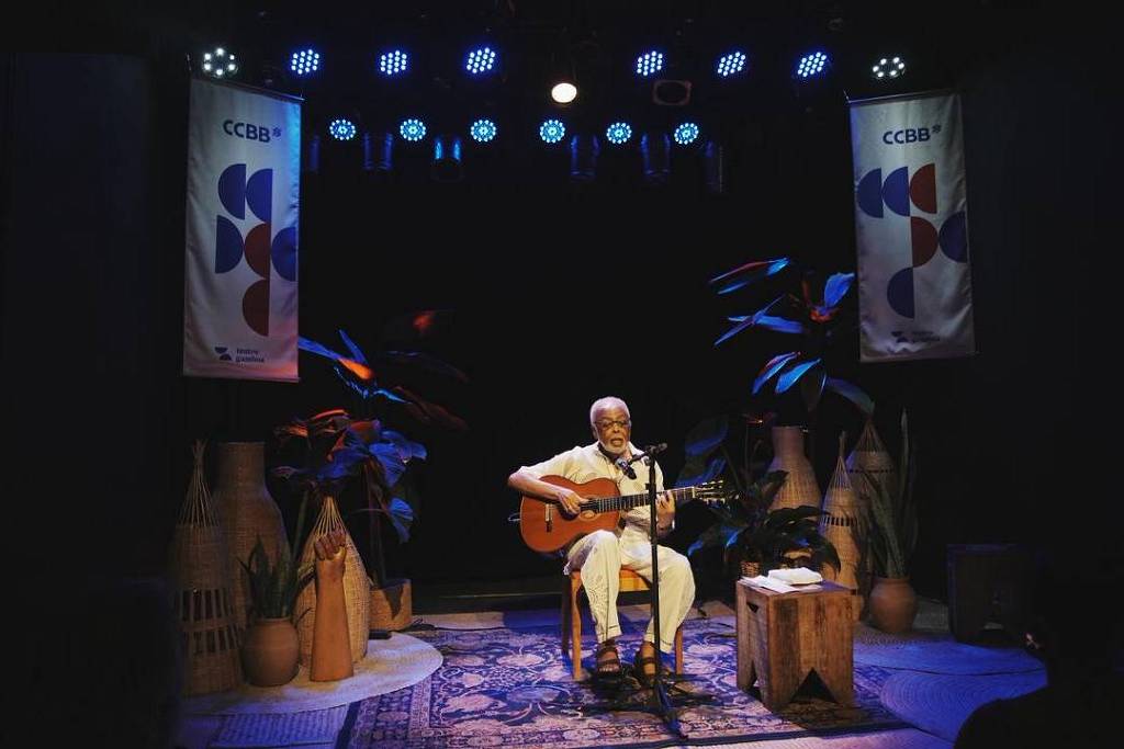 Músico sentado no centro do palco toca violão sob luzes azuis. Dois banners do CCBB pendem ao fundo, com plantas e objetos decorativos ao redor.