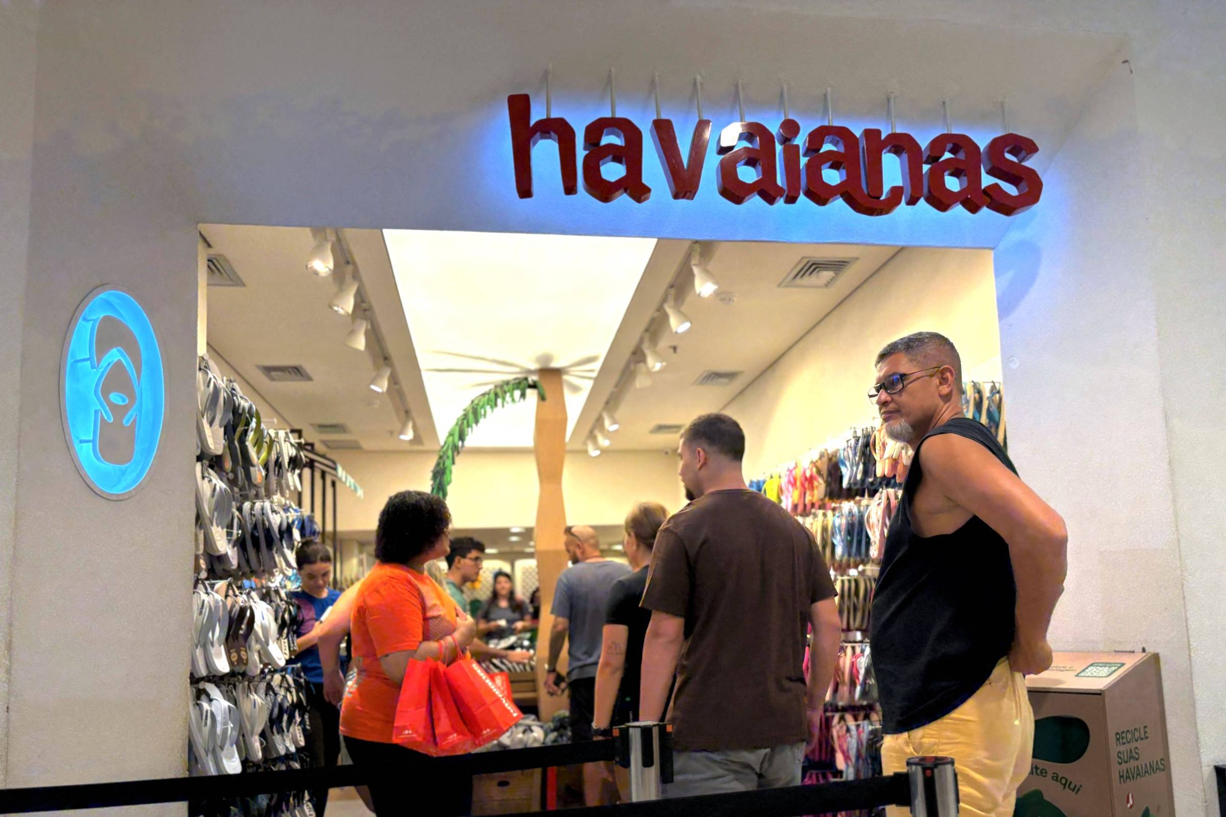 Clientes aguardam em fila na entrada de loja Havaianas em shopping. Paredes internas exibem diversas sandálias penduradas. Iluminação clara destaca produtos e ambiente.