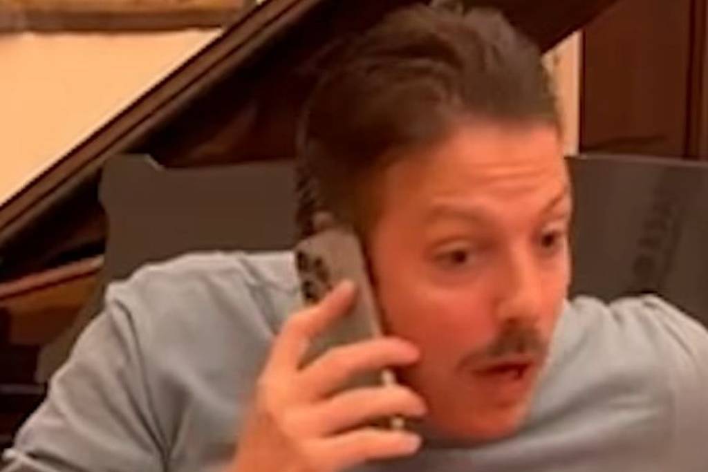 Homem de camiseta azul claro fala ao telefone sentado em frente a um piano de cauda preto. Texto na tela diz: