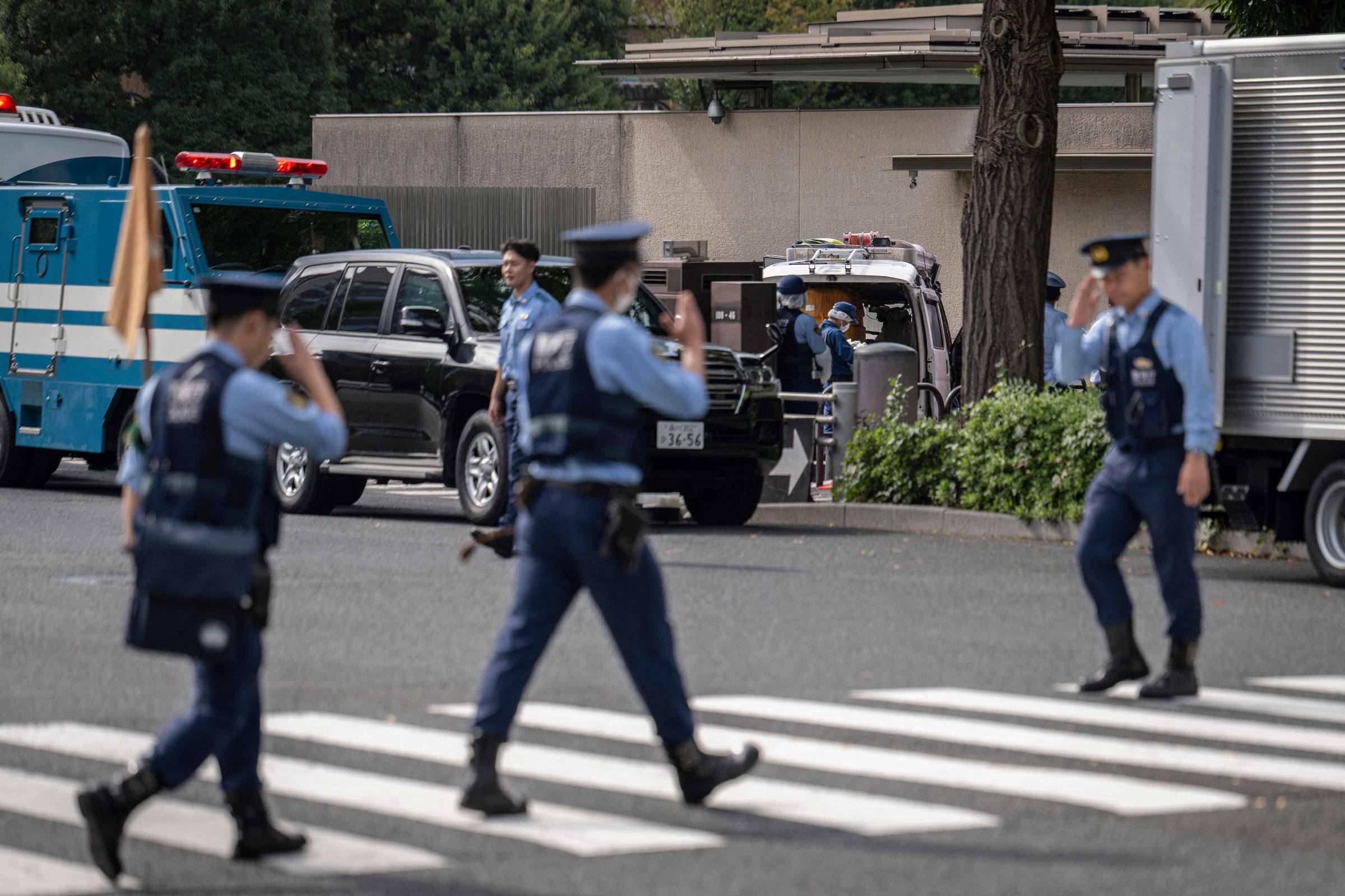 Três policiais em uniforme azul cruzam uma faixa de pedestres em frente a veículos oficiais estacionados, incluindo uma van e um carro preto. Um homem em roupa clara está próximo à van, e há árvores e um muro ao fundo.