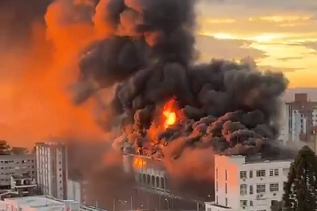 Fogo intenso consome o topo de um prédio em área urbana, com grandes nuvens de fumaça preta e alaranjada se espalhando pelo céu ao pôr do sol. Edifícios vizinhos e árvores estão visíveis ao redor.