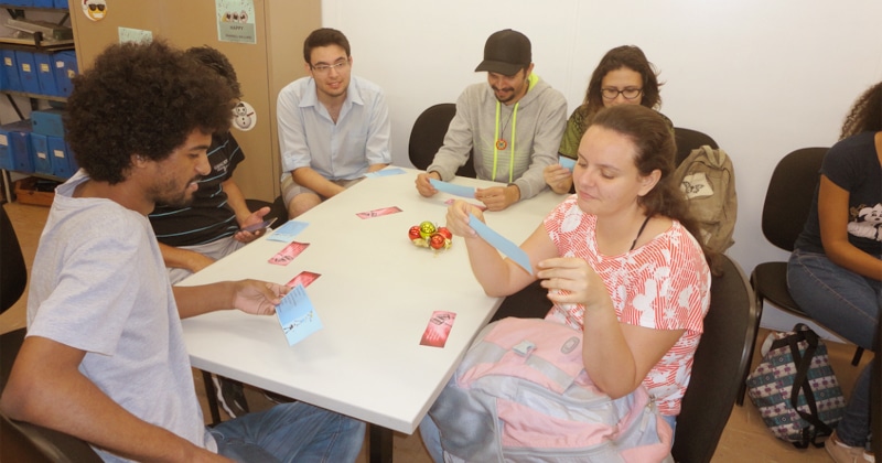 Seis pessoas sentadas em volta de uma mesa pegando papéis numa atividade educativa