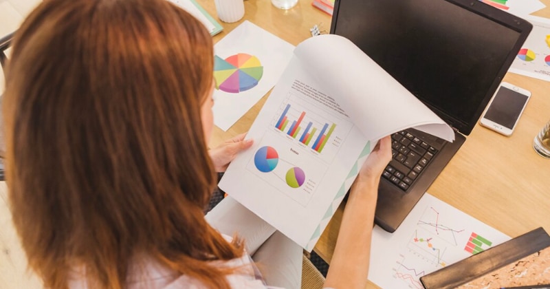 Mulher de jaleco branco de costas olhando para um papel com gráficos e à frente uma mesa com notebook